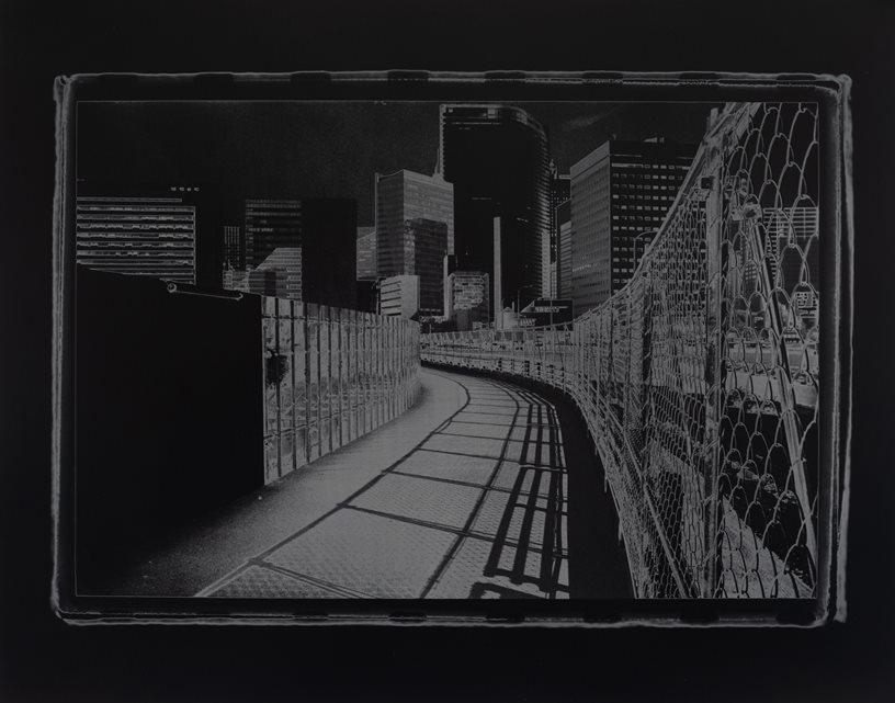 photograph by Larry Gawel - Tsukiji No More, clockwise rotation, gelatin silver print, which shows a walkway curving to the right, with a fence on the left and a wall on the right that follows the same curvature. Visible in the distance are several tall buildings, possibly indicating a downtown metropolitan vantage point.